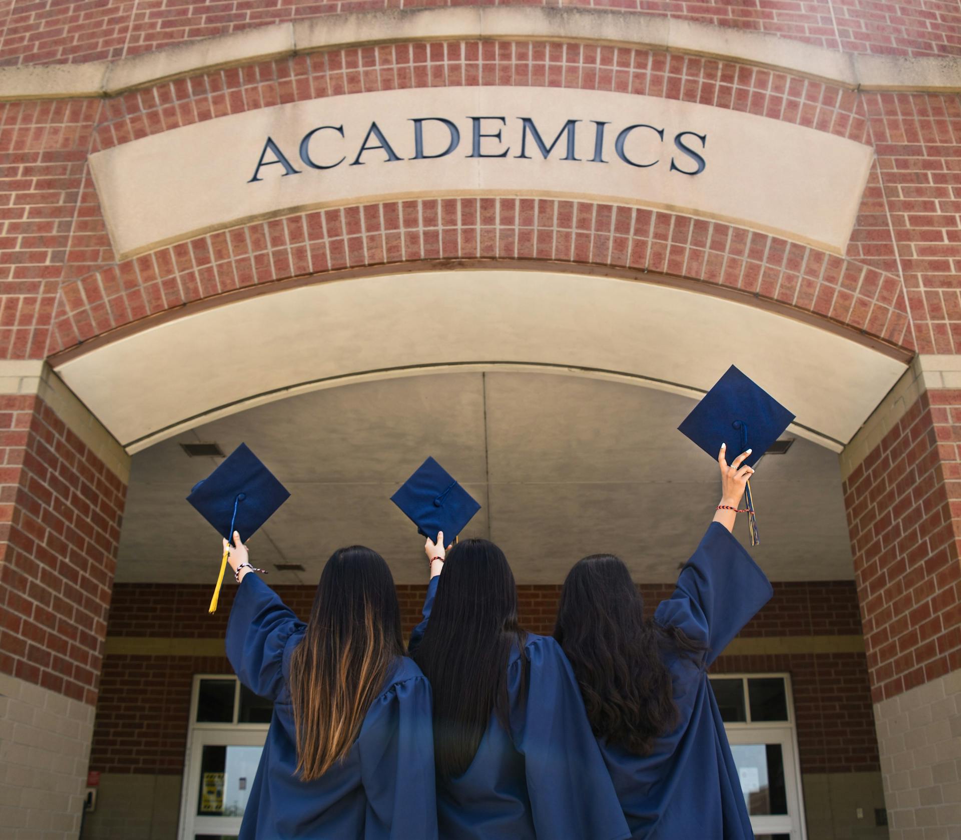 Three women are standing backwards and holding mortarboard hats during graduation. 